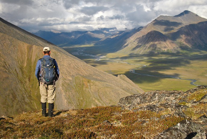 A hiker overlooks a mountain valley as clouds create shadows across the landscape A hiker overlooks a mountain valley as clouds create shadows across the landscape