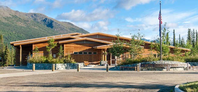 View of the entrance area of the Arctic Interagency Visitor Center with gravel praking lot, wooden stanchions, and cement sidewalks