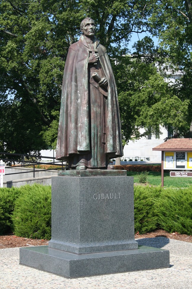 A color photo of a bronze statue of Father Pierre Gibault standing atop a square black stone base.