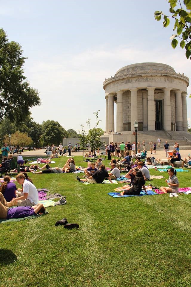 Adults and children sit on towels and blankets on a green lawn in front of the George Rogers Clark Memorial building.