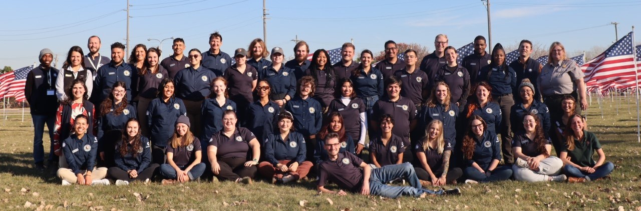 lt= A group photo of Community Volunteer Ambassadors members posing in front of a USA flags while wearing navy blue polos with the official Community Volunteer Ambassador logo.