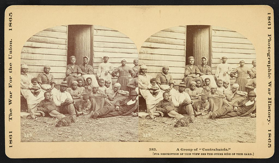 A black and white photo of former slaves, or contraband, sit around the door opening of a structure. There are around twenty men and women including one infant.