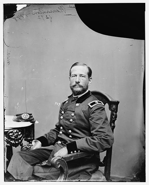 A black and white photo of a Union officer. He sits in a chair wearing his uniform and he has a beard and a mustache. His hat sits on a table to his right.