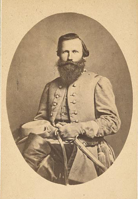 A black and white photo of a Confederate soldier sitting in a chair. He is wearing his full dress uniform and is holding his hat and saber in his lap. He has a long and full beard and mustache.