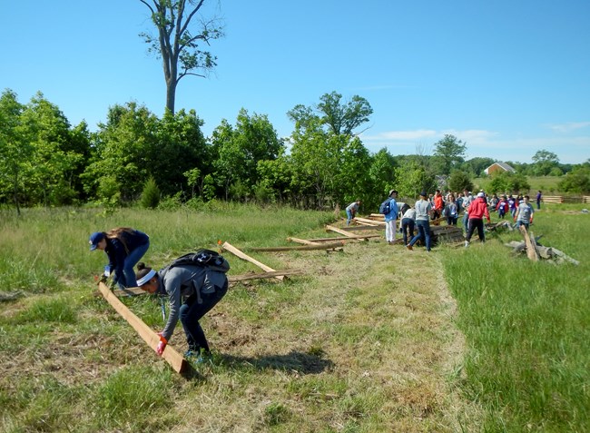 A group of people begin to build a wooden fence in the middle of a mowed green grass field. The wooden fence has been laid one course in a zig-zag pattern. Two people are laying one piece of fence near the camera while a large group readies more.