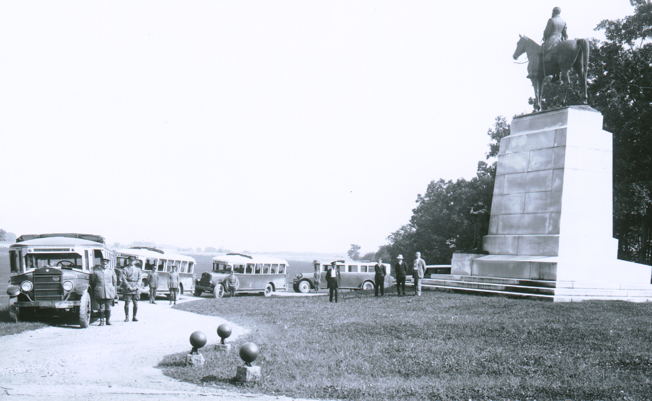 LBGs at VA Memorial late 1920s-30s A black and white photograph of a tall white monument with a horse and rider on top is on the right. There is a curved dirt roadway on the left with four buses and their drivers parked and standing along the road.