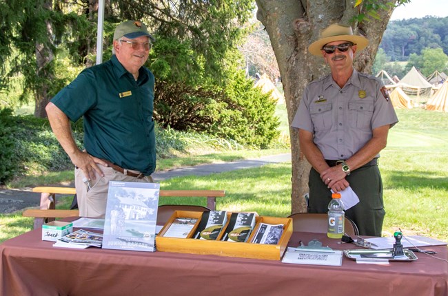 Two men stand behind a display table with a brown table cover. The man on the left is dressed in a short sleeved green shirt, khaki pants, and green ball cap. The man on the right is dressed in a short sleeved grey buttoned up shirt, green pants, and hat.