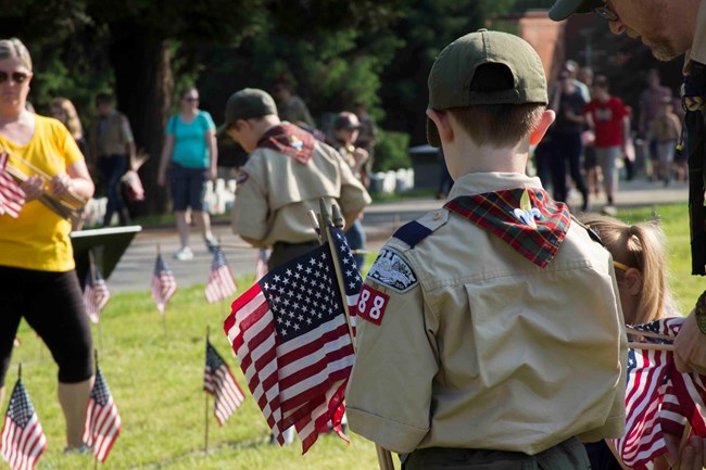 A young boy in a scout uniform, a short sleeved tan shirt and green ball cap, carries small United States flags in a national cemetery. Many other scouts and people are helping to plant flags throughout the cemetery.