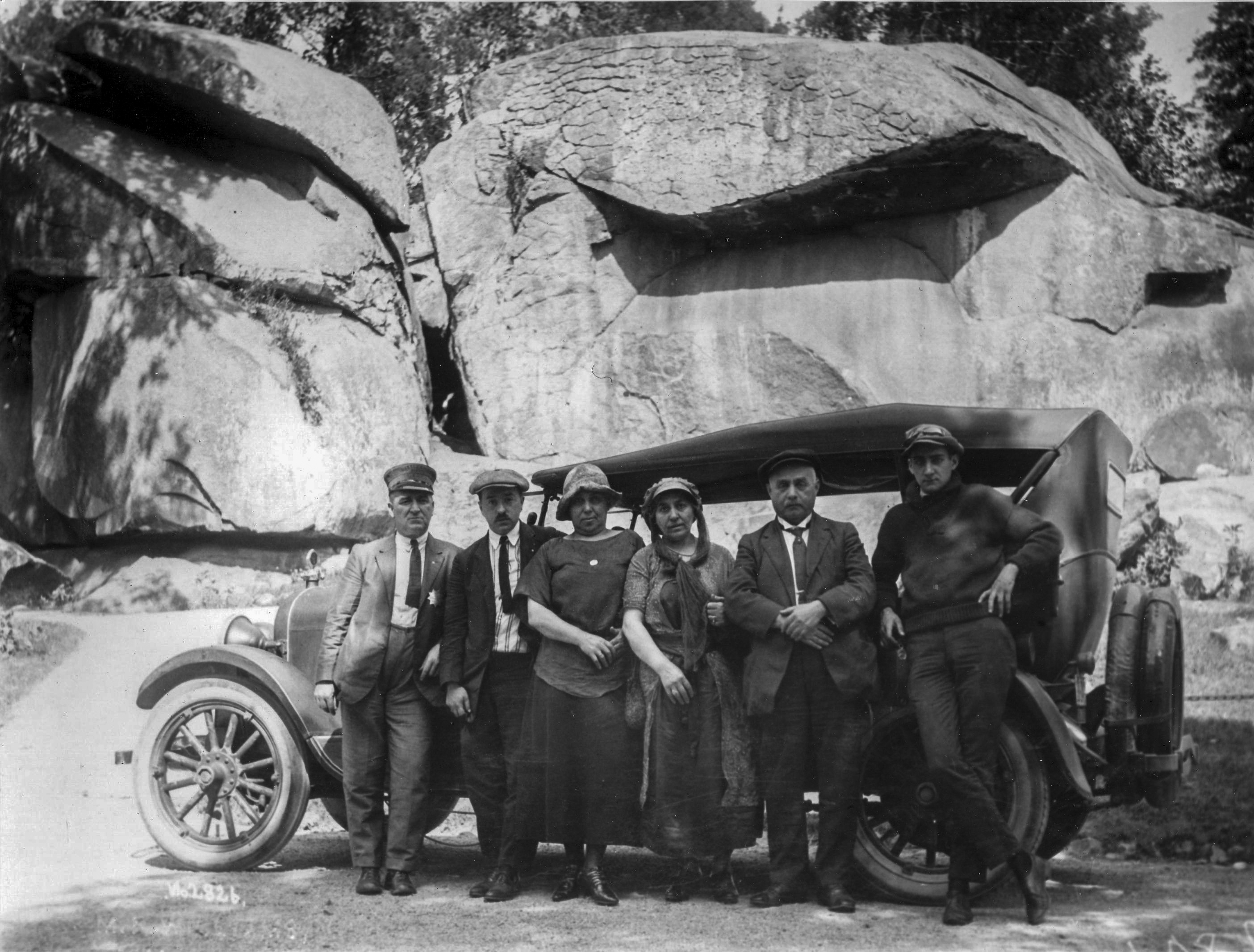 An unknown Licensed Battlefield Guide with a group of visitors in Devil's Den. The photograph was taken circa 1920. A black and white photo of a group of six adults standing in front of an 1920s era automobile. Behind the group and automobile is a wall of very large boulders.