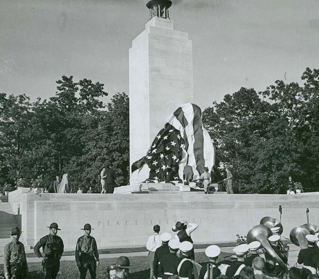 A black and white photo of a band and crowd in front of a large white stone monument. A large flag is seen falling from the top of the monument.