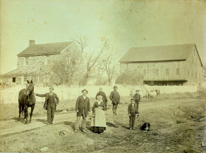 Basil Biggs and Wife Mary stand in front of their stone home along a dirt road. A barn is in background, and several other individuals are also in photograph.