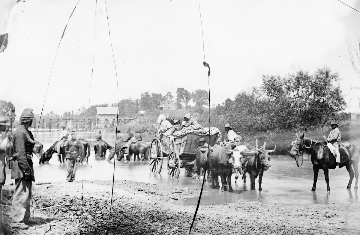Historical black and white photograph of a group of Black people, including men on horses and a woman on a wagon pulled by oxen crossing a low river while watched by white US soldier.