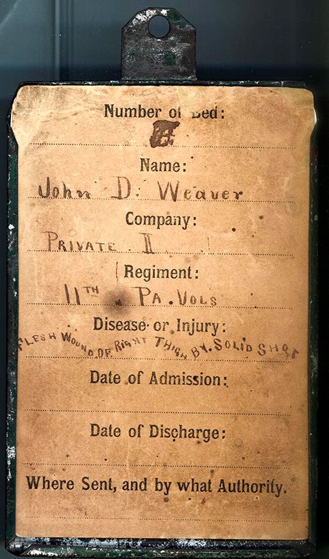 Paper hospital bed tag mounted on a rectangular tin sleeve. Information on the tag includes: John D. Weaver/ Private II/ 11th Pa Vols./ Disease of the flesh, wound right thigh, by solid shot. John D. Weaver enlisted as a Private for 3 years in Co. I