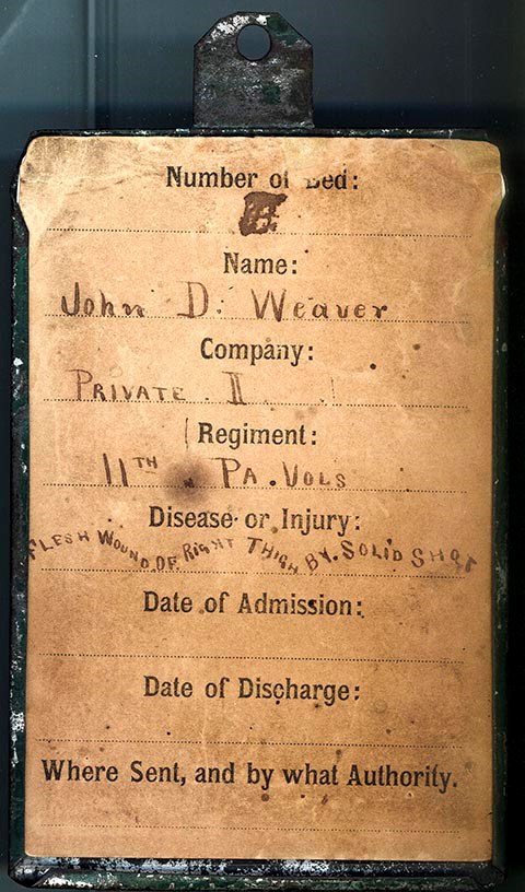Paper hospital bed tag mounted on a rectangular tin sleeve. Information on the tag includes: John D. Weaver/ Private II/ 11th Pa Vols./ Disease of the flesh, wound right thigh, by solid shot. John D. Weaver enlisted as a Private for 3 years in Co. I