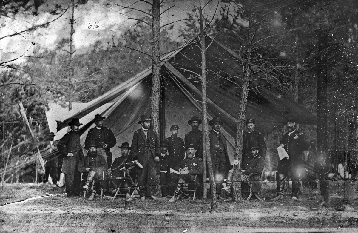 Historical black and white photograph of US General Ulysses S. Grant, standing, with his military staff, all seated in front of a tent.