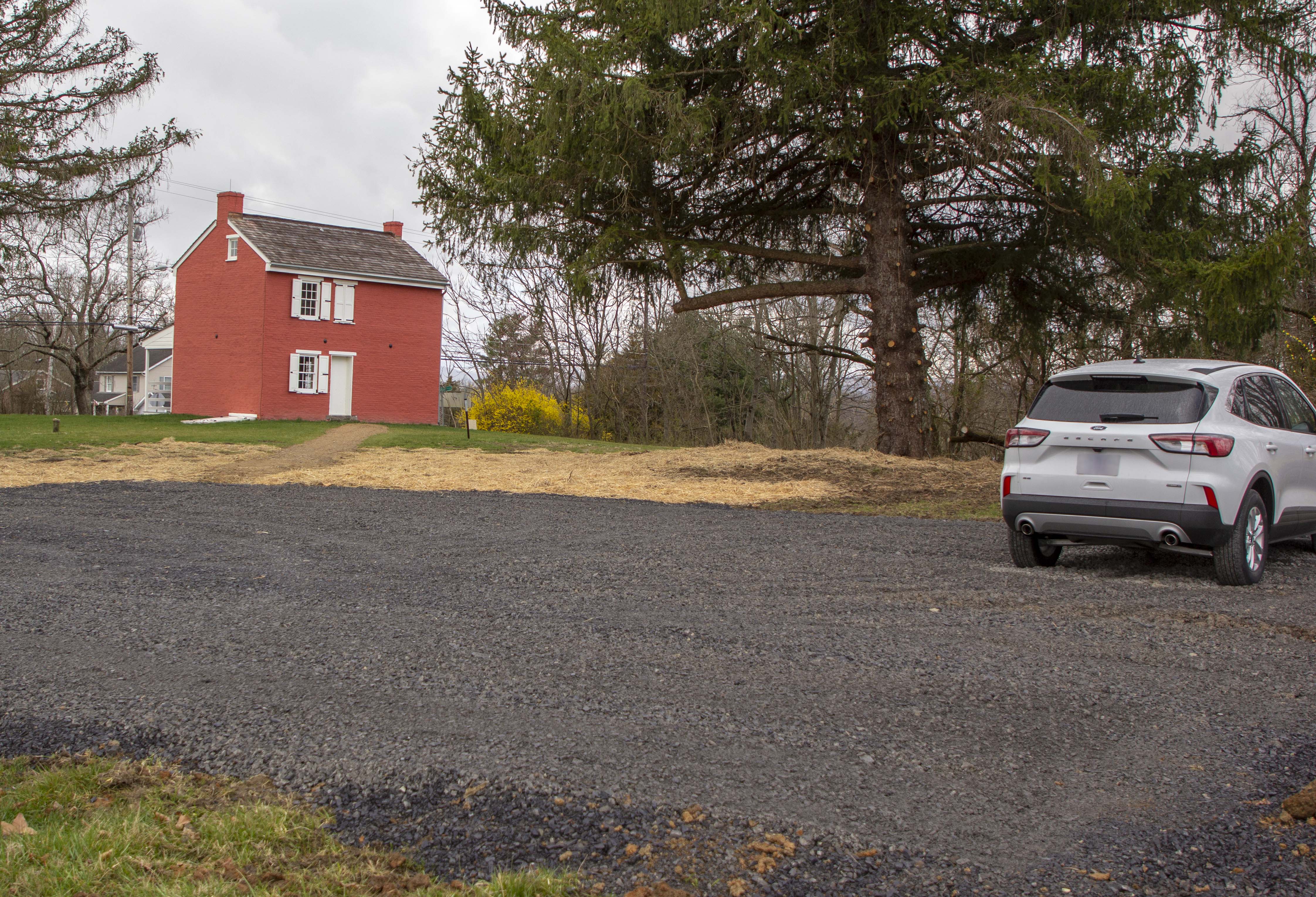 In the foreground is a dark gravel parking lot with a small white SUV on the right. In the distance in the center is a large pine tree and on the left is a small, two story, red brick house with white shutters and door.
