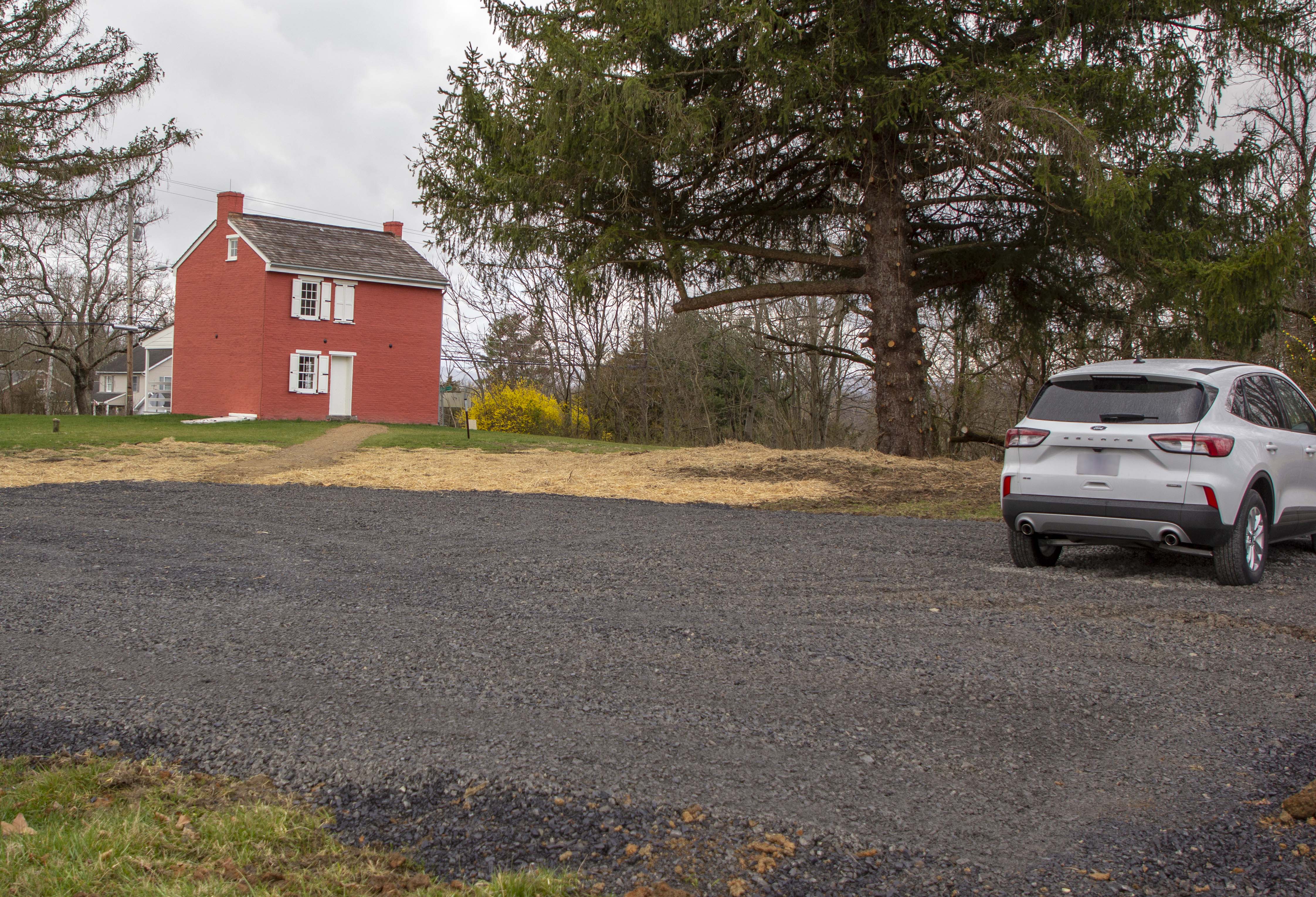 Wisler house parking In the foreground is a dark gravel parking lot with a small white SUV on the right. In the distance in the center is a large pine tree and on the left is a small, two story, red brick house with white shutters and door.
