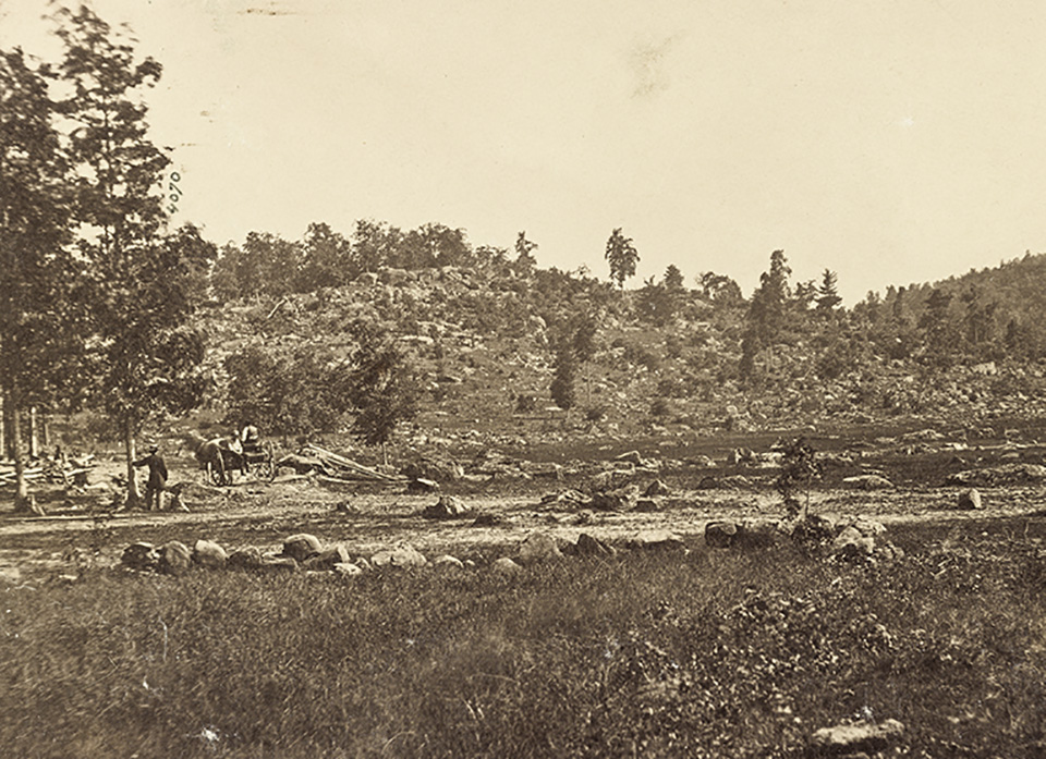A black and white photograph, taken in 1863, of a rocky hill in the distance. There is a man standing against a tree in the left center and his horse and buggy sit nearby.