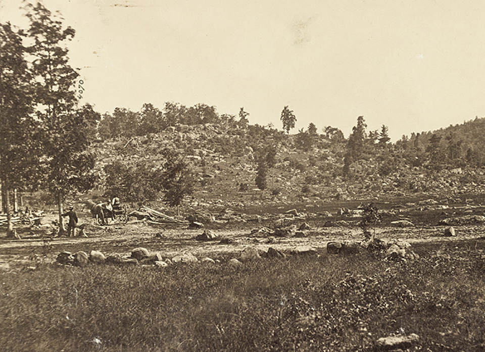 Little Round Top from the Valley of Death A black and white photograph, taken in 1863, of a rocky hill in the distance. There is a man standing against a tree in the left center and his horse and buggy sit nearby.