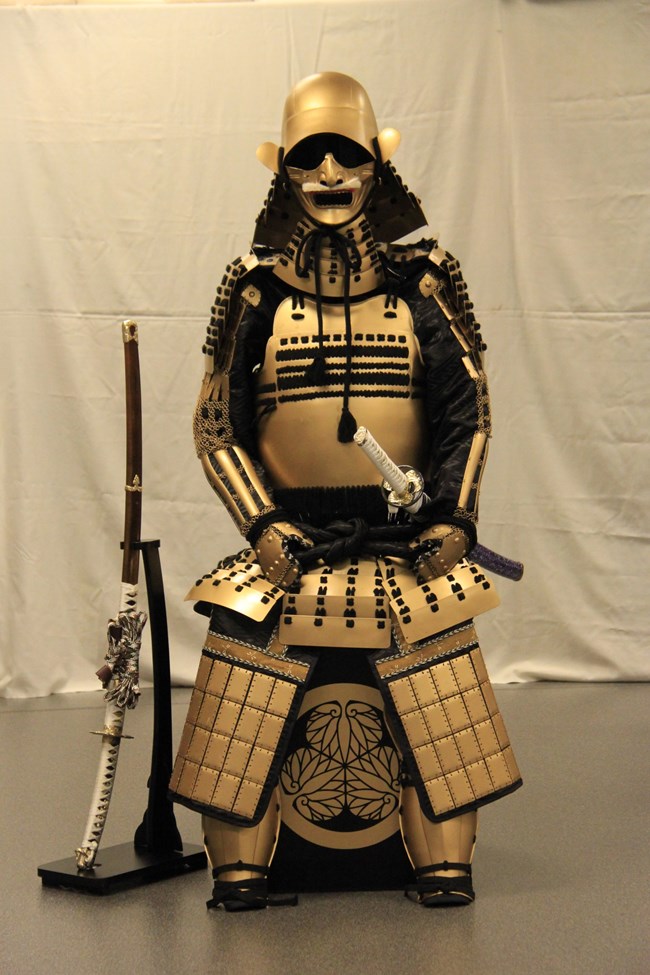Traditional Samurai armor with a katana on the left side hip and another on a stand to the right.