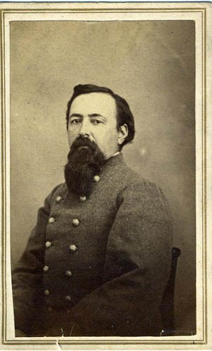 Black and white photo of a man, seated, with a black beard and black hair wearing a gray military coat