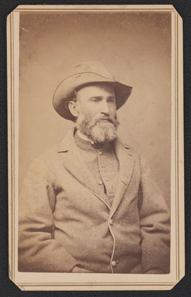 Black and white photograph of a bearded man wearing a gray jacket over a gray vest and a gray hat