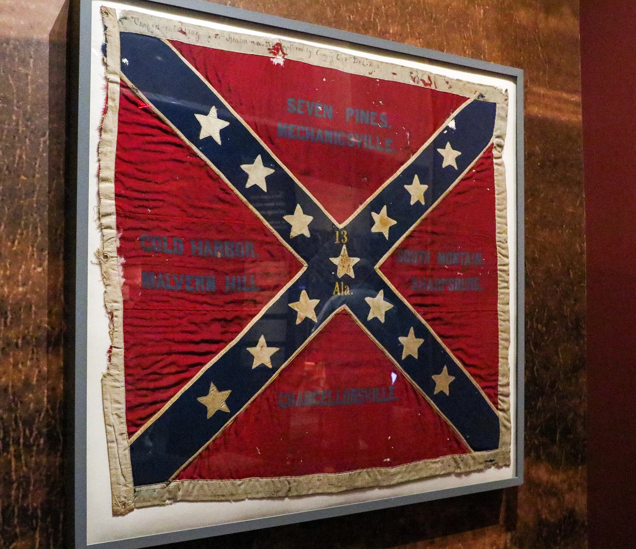 13th Alabama Battle Flag A Confederate battle flag hangs on a wall. The flag has a red background with diagonal blue lines and thirteen white stars on the blue lines.