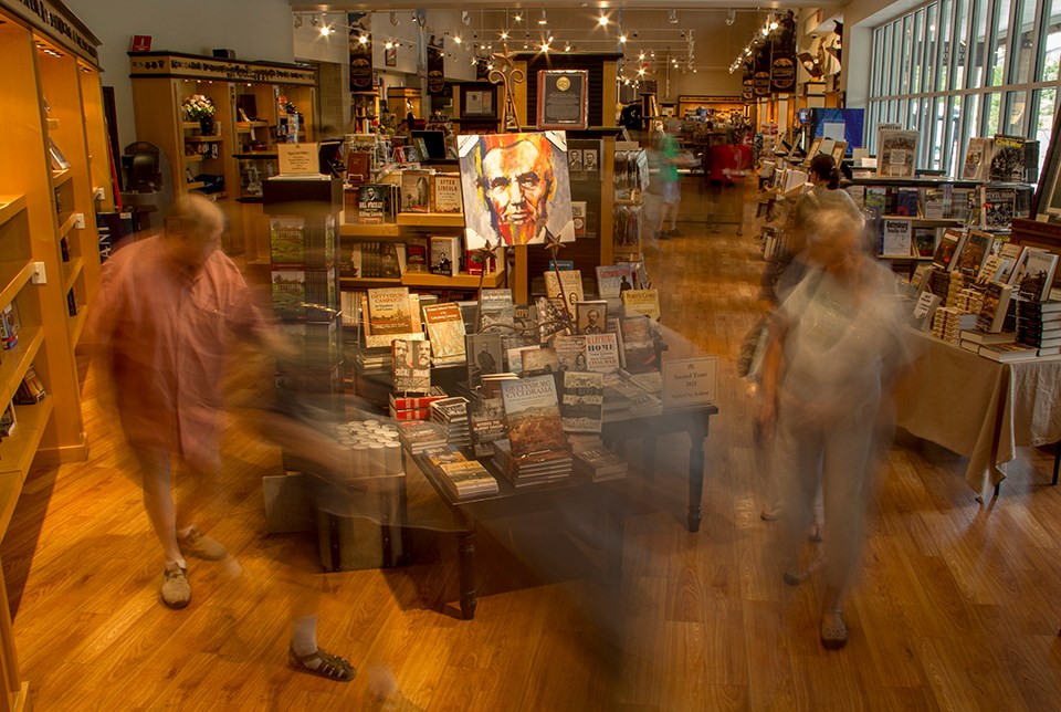 Visitors shown moving through the park bookstore. They are walking around tables of book displays and bookcases line the walls.