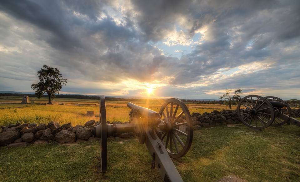 The sun sets over the battlefield. Two cannons sit in front of a stone wall and a monument and a tree are in the left distance.