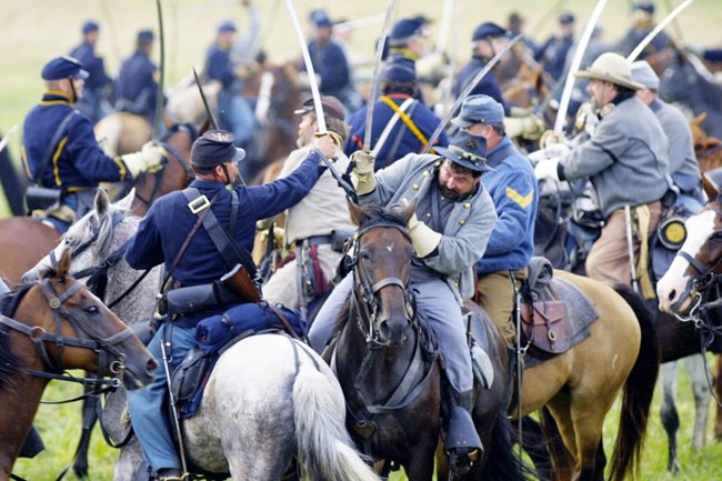 A scene of cavalry soldiers reenacting a battle. There are many soldiers in blue and in grey uniforms, some holding swords in the air.