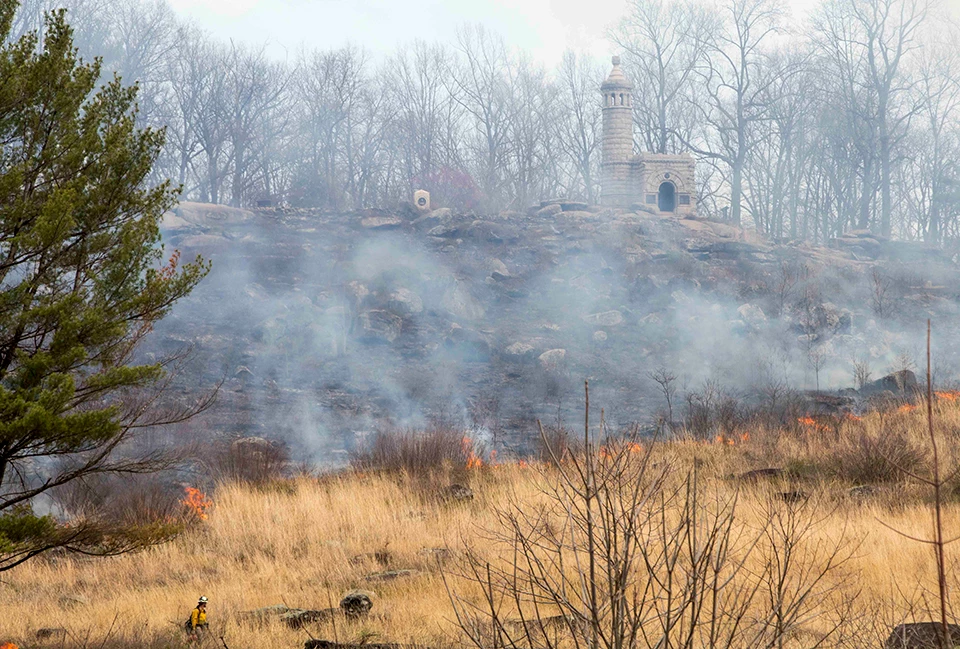 A prescribed fire on Little Round Top slowly moves from left to right down the slope of the hill. A prescribed fire on Little Round Top slowly moves from left to right down the slope of the hill. There are monuments along the top of the hill. The fire is moving through brush.