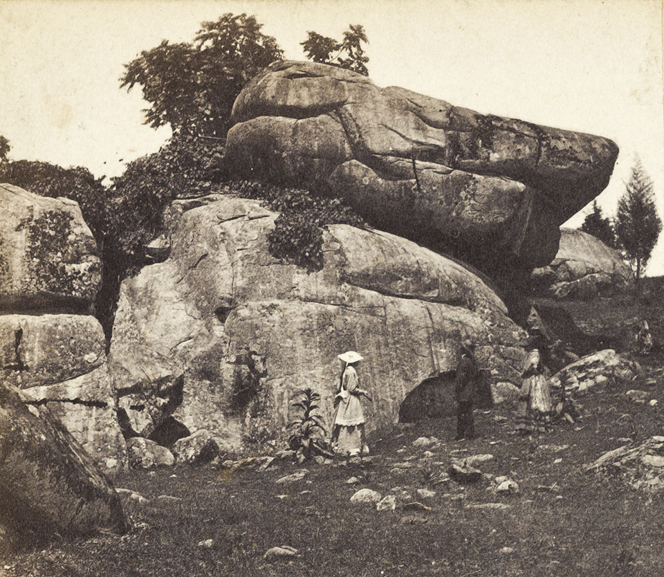 Unidentified visitors to Devil's Den. Photo taken between 1863-1880. A black and white photograph of two women and one man standing in front of a stack of very large boulders that tower over their heads.