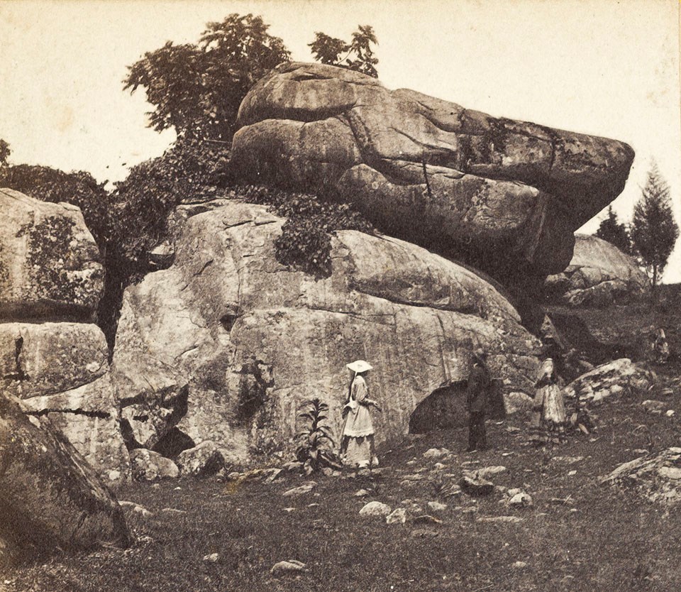 A black and white photograph of two women and one man standing in front of a stack of very large boulders that tower over their heads.