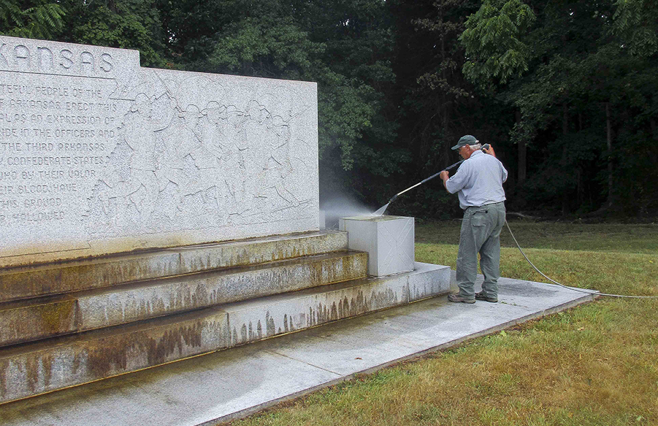 Arkansas Memorial cleaning A park employee uses a pressure washer to clean the Arkansas Memorial. There is an etched wall of soldiers and three steps of light colored granite.