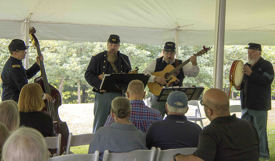 The Libby Prison Minstrels at Gettysburg during the 2017 Music Muster. Four musicians, dressed in blue Union uniforms, play four different instruments to a crowd of people under a white tent.