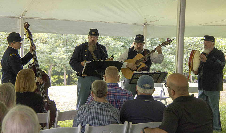 Four musicians, dressed in blue Union uniforms, play four different instruments to a crowd of people under a white tent.