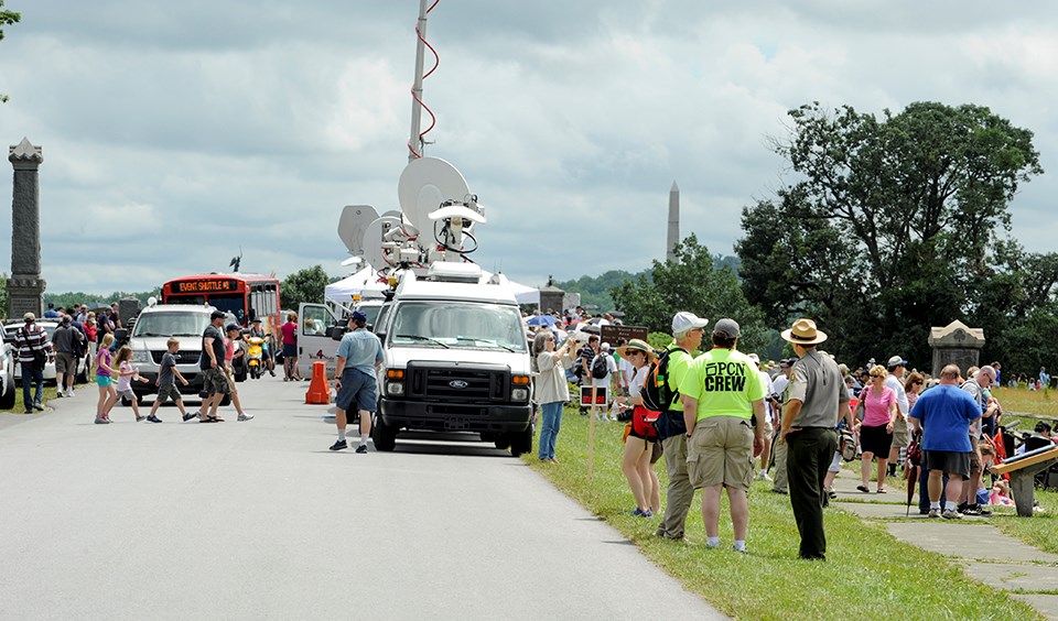 Press trucks are parked along the side of the road. A park ranger talks to visitors in the foreground and lots of visitors walk around the battlefield.