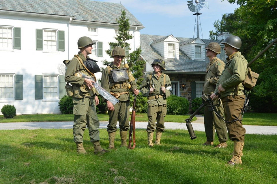 Members of the Furious Fourth living history group stand in front of the Eisenhower home. A group of five men, dressed in World War two uniforms and carrying proper time period reproduction weapons and equipment, stand in front of white sided house with green shutters and a small stone addition. There is a windmill in the right background.