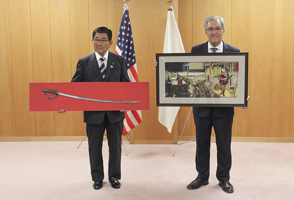 Two men stand in front of a light brown paneled wall and the American and Japanese flags. The Japanese man on the left holds a Civil War saber mounted on a red panel. The American man on the right holds a piece of art in a black frame.