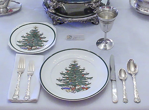 A dining room place setting of two china plates, two silver forks, two silver spoons, one silver knife, and one silver goblet. The plates are white with a green ring around the edge and a decorated Christmas tree with presents underneath in the center.