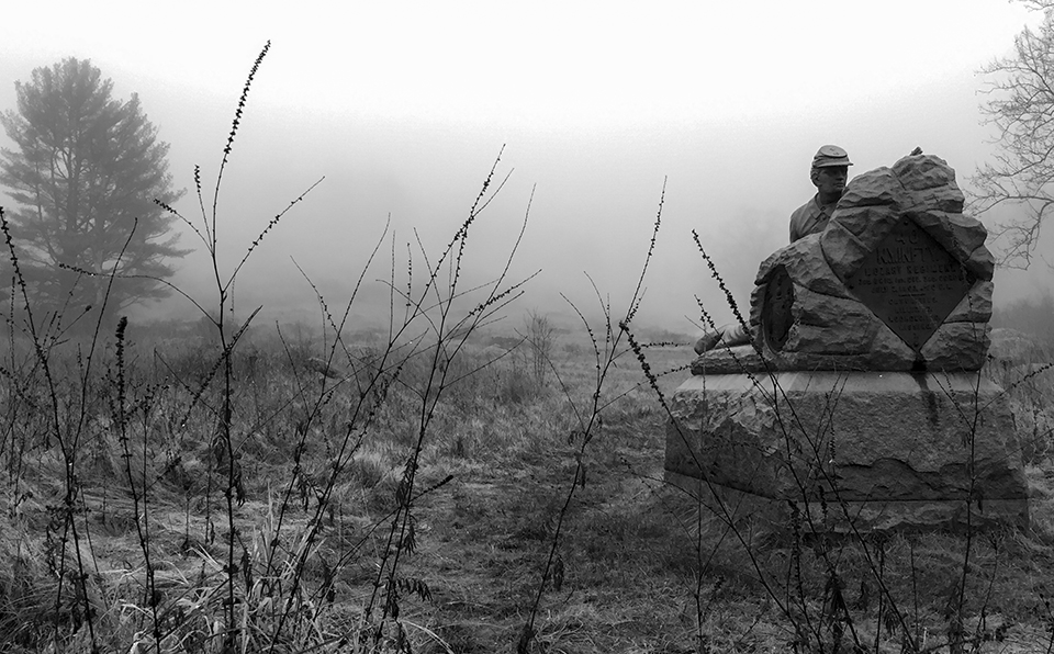 A black and white photo of a meadow with a monument on the right side depicting a Union soldier behind a stone wall ready to fire his rifle.