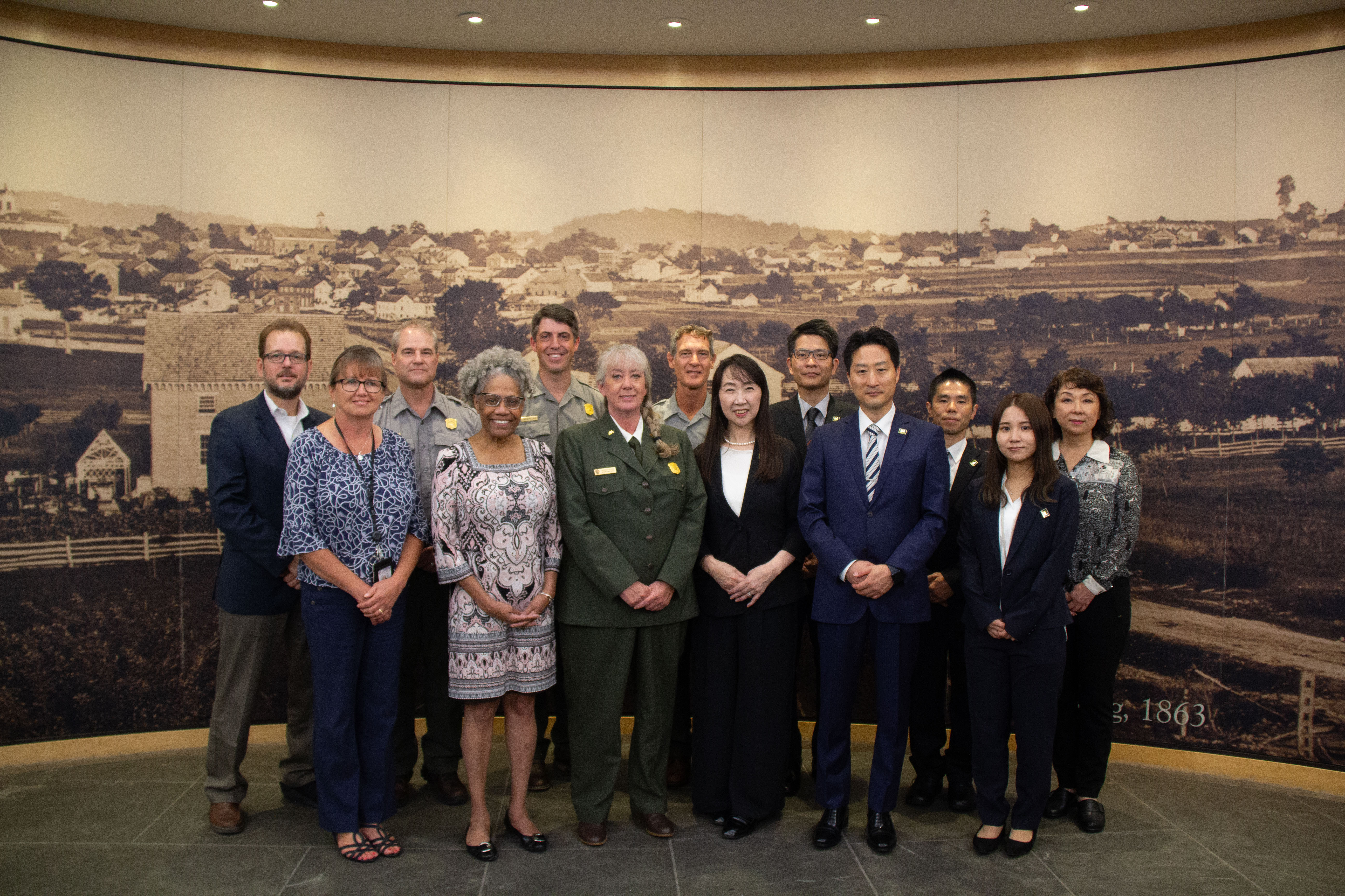 The delegates from Gifu Prefecture, Sekigahara, Japan stand with Gettysburg National Military Park and Gettysburg Foundation staff inside the Museum and Visitor Center. A group of thirteen men and women stand for a photograph in front of a large mural backdrop that depicts Gettysburg, Pennsylvania in 1863.