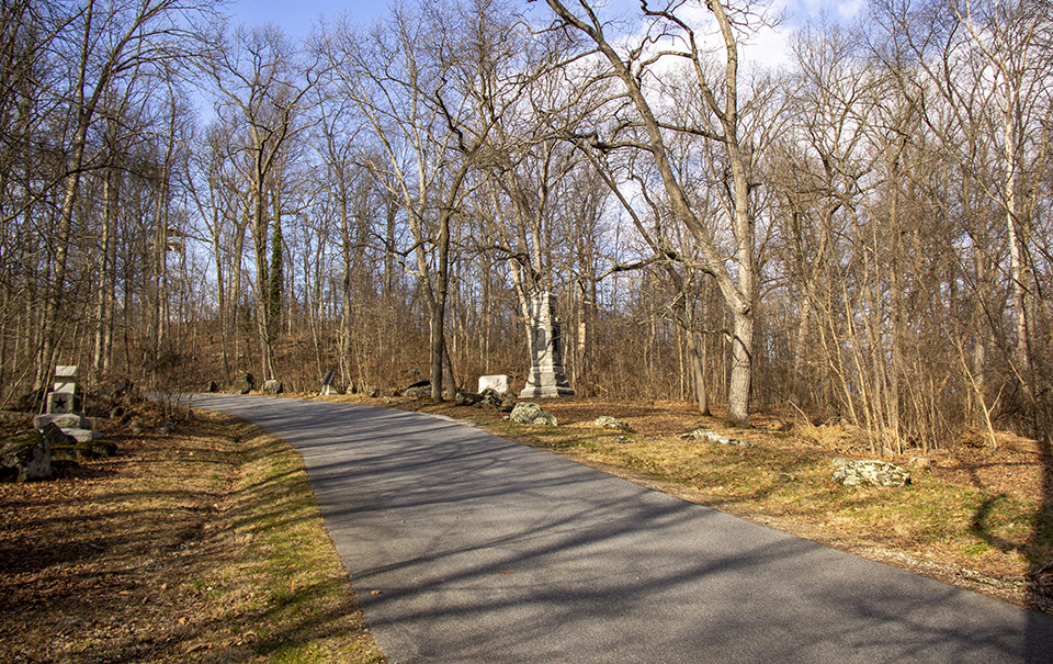 A paved road runs up a hill and bends away to the left. One granite monuments sits on the left of the road and two more sit on the right of the road. The area is heavily wooded but the trees are bare.