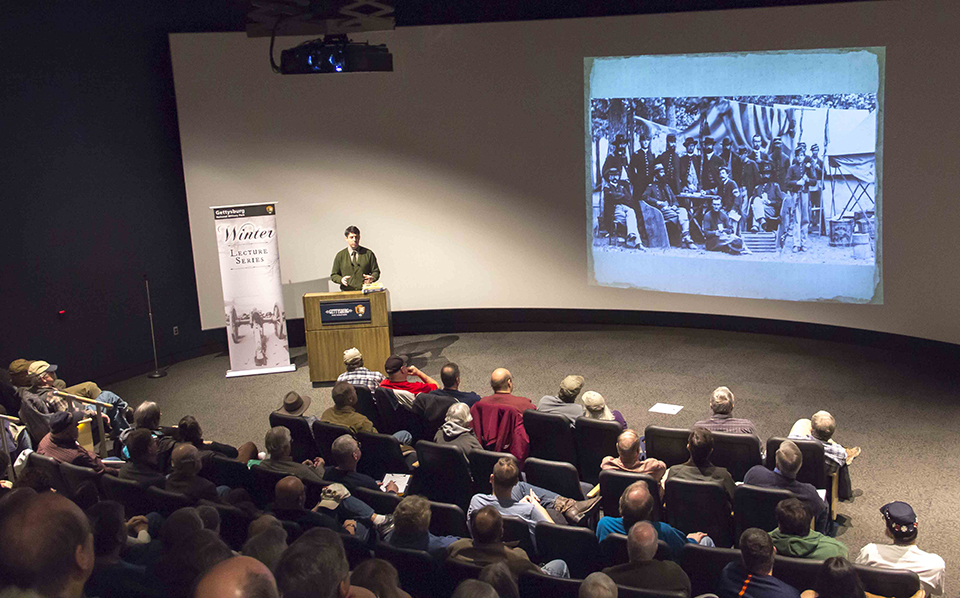 A park ranger presents a program during the Winter Lecture Series. A crowd of people sit in an amphitheater as the ranger shows different slides on the screen.