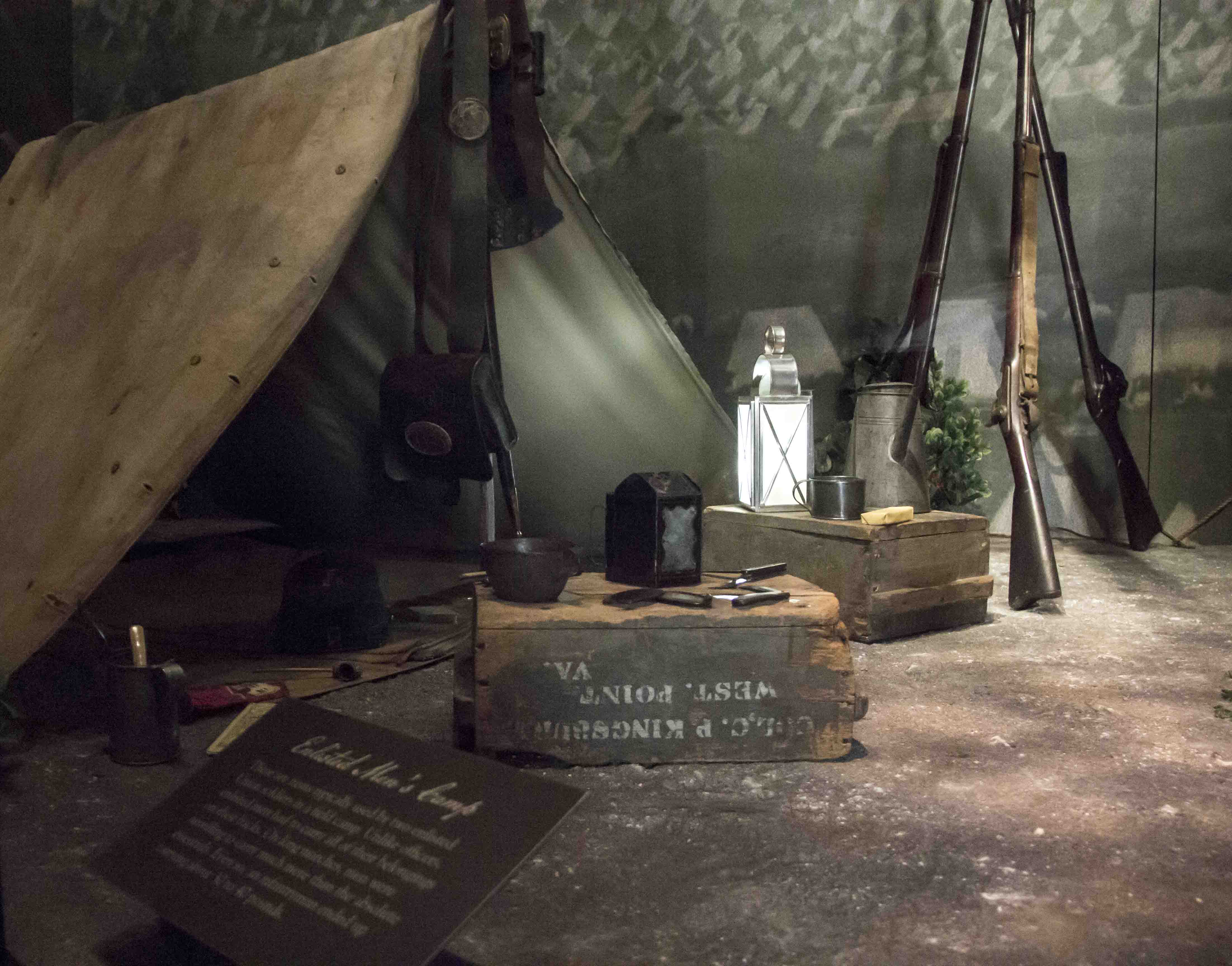 A museum exhibit of camp life depicts a Civil War soldier's tent, lantern, guns, tin cup, tin coffee pot, two wooden boxes, and an interpretive panel.