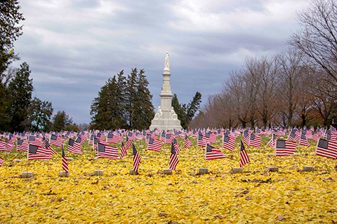 A tall white granite monument stands in the center in the distance and the foreground is covered by small American flags and yellow fall leaves.