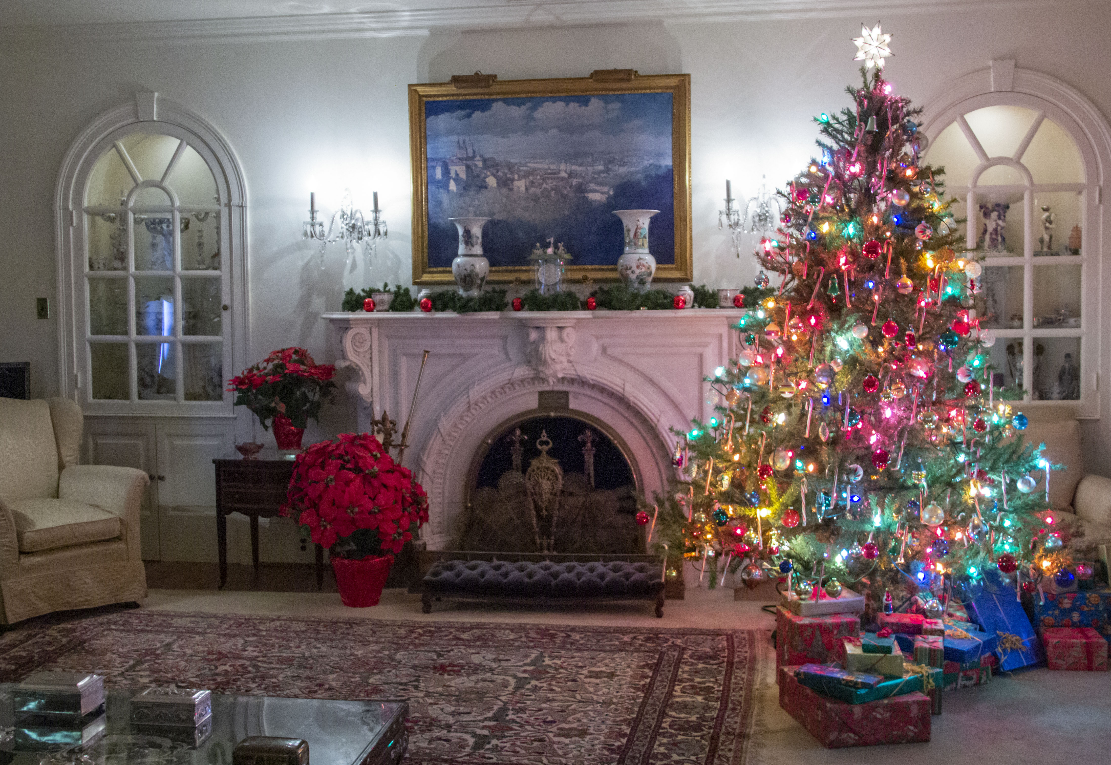 A decorated room for Christmas. There is a colorful lit tree with wrapped presents on the right. There is a fireplace and mantle with a painting in the middle. There is a tan armchair and a red poinsettia on the right.