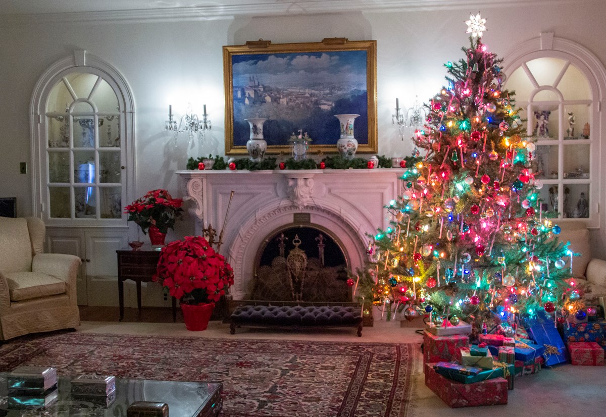 A decorated room for Christmas. There is a colorful lit tree with wrapped presents on the right. There is a fireplace and mantle with a painting in the middle. There is a tan armchair and a red poinsettia on the right.