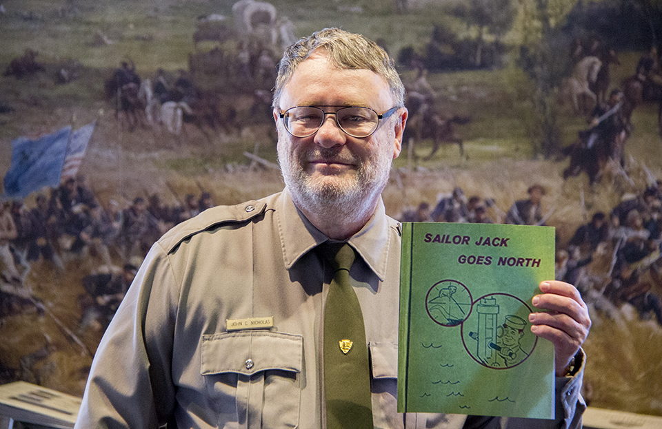 A park ranger holds the book, "Sailor Jack Goes North,". He is in his park uniform, holding the green book, in front of the park information desk.