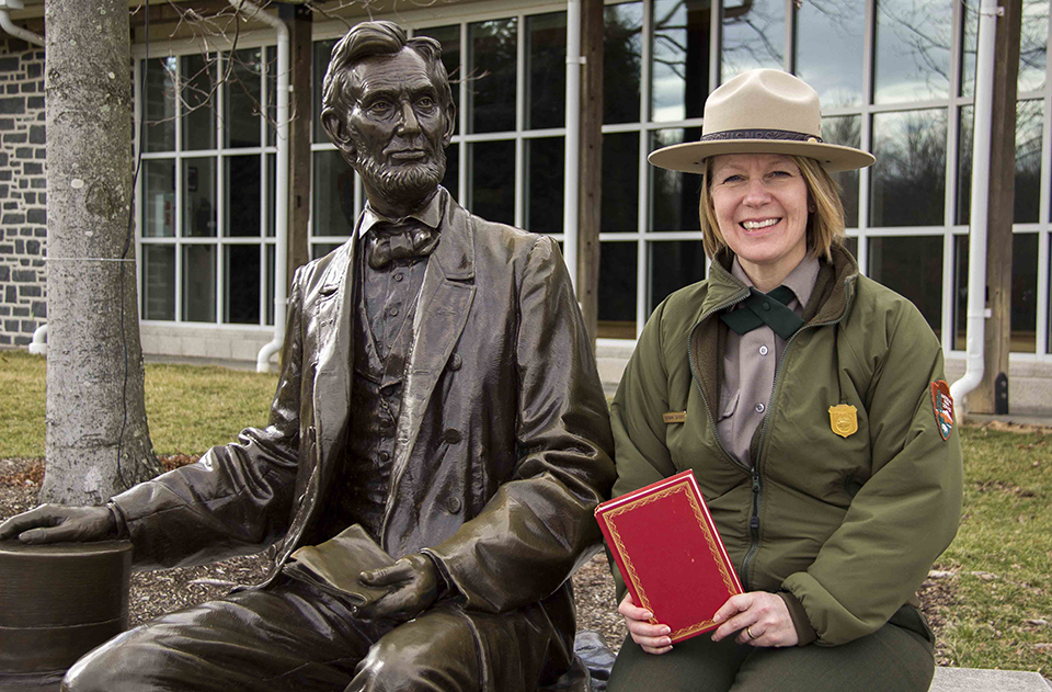 Barb Sanders sits next to a statue of Abraham Lincoln. She is holding a small red book.